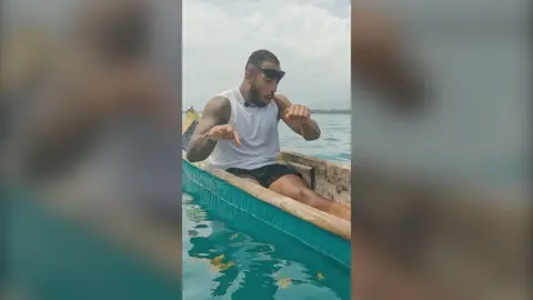 Race Across The World contestant Tyler West in a narrow fishing boat looking shocked after catching a fish. He's holding a fishing line in his left hand and moving his right hand away and wearing sunglasses on his head