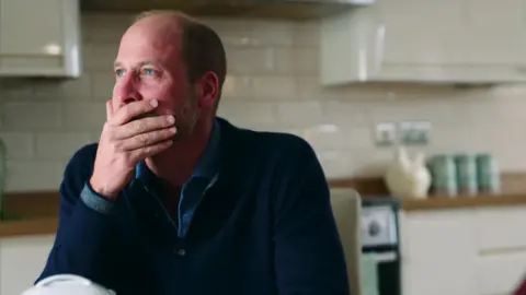 Prince William of Kensington Palace covers his mouth with his hand, wearing a navy blue sweater in a white-tiled kitchen 