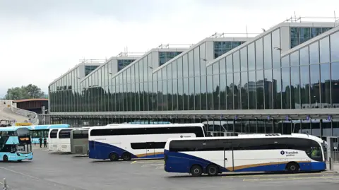 Pacemaker Translink buses are lined up outside the station. An airport express bus is arriving. The building is surrounded by windows and the roof is a wave shape.