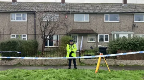 A street with blue and white police tape in front of it. A police officer stands looking at the camera with his hands clasped.