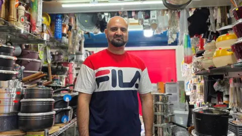 Paul Moseley/BBC Mohammed is wearing a blue, white and red 'Fila' t-shirt. He's standing in the middle of the stall, surrounded by cookware.