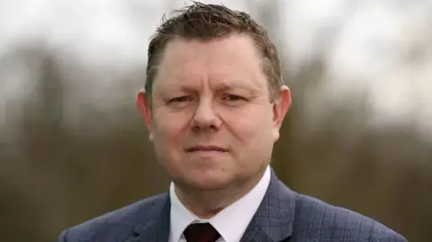 PA Media John Apter looks at the camera. He is wearing a checked suit, white shirt and tie.