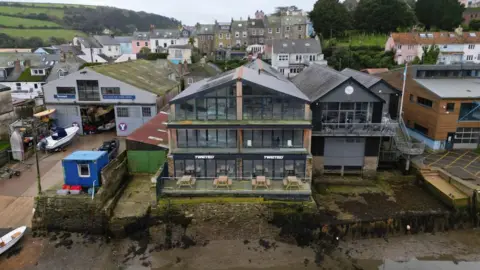 A three storey building overlooking an inlet with a boatyard to one side and another building to the other with tables and chairs on the ground floor and houses behind.