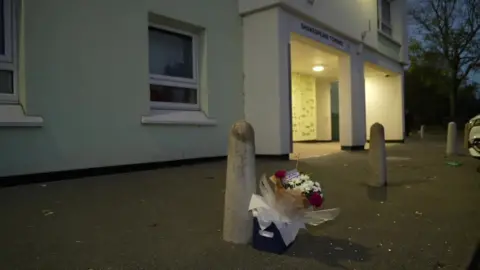 A bunch of flowers sits up against a bollard near the entrance of a tower block, with a sign above the doorway reading "Shakespeare Towers". The sky is getting dark, with a bright light shining at the entrance to the block. 