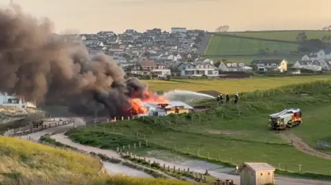 A beachside building is on fire with large plumes of black smoke coming away from it and over the beach. Fire crews are seen spraying the property with water.