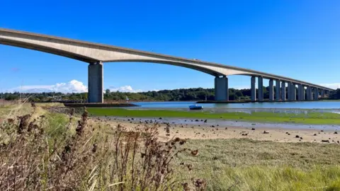 The Orwell Bridge - a 1km (0.6-mile) long concrete bridge over the river Orwell - seen from the riverbank below. There is grassland, a small boat and water. It is a sunny day.
