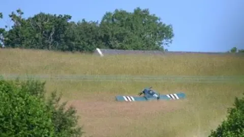 Field surrounded by trees with a light aircraft in the grass.