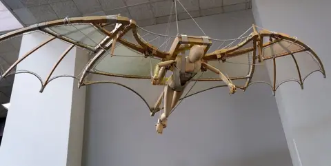 Getty Images A model of Leonardo Da Vinci's wooden flying machine suspended from a white tiled ceiling.