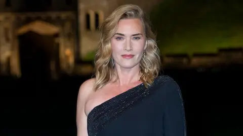 Getty Images Kate smiles while looking at the camera. She is wearing a off the shoulder black sparkly dress and has blonde curly hair.