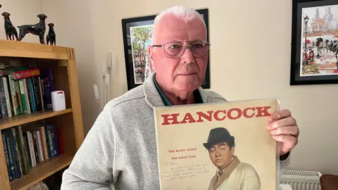 Alan Florence, has very short cropped grey hair he has silver framed glasses and is wearing a beige woollen cardigan with a green shirt beneath with the top buttons undone, behind him is a bookcase with vinyl albums and books and a small telescope in front of it.