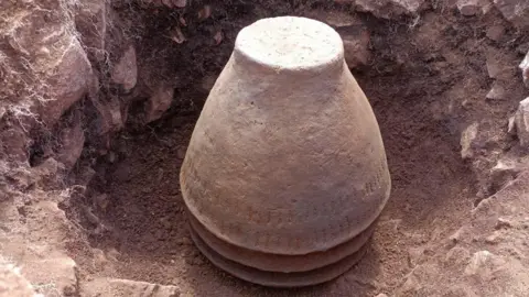 NNP Large red-brown pot which looks like a flower pot in a hole in the ground. It looks like it is upturned.