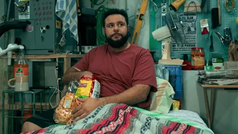 Eddy Chen/Netflix Mo in the show, wearing a red t-shirt, sitting in bed and eating crisps. Around him are tools and a fan as he looks off into the distance.