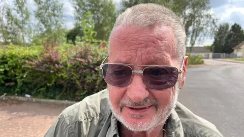 David Buchanan has short grey/brownish cropped hair and a matching short beard with moustache. He is wearing dark glasses and khaki shirt, behind him is a roadway and some hedges and trees.