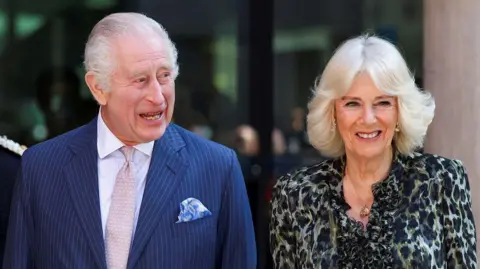 Reuters King Charles III and Queen Camilla. He is wearing a blue pin-striped suit and has a pink tie and white shirt. She is wearing a tortoise shell blouse. They are both smiling.