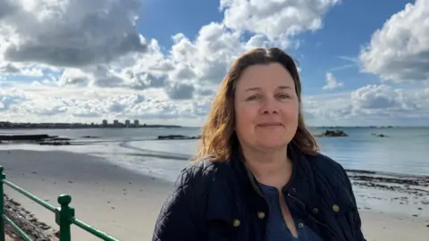 Fiona Kerley pictured in Havre De Pas. The beach and sea is behind her. She is wearing a black jacket and blue top.