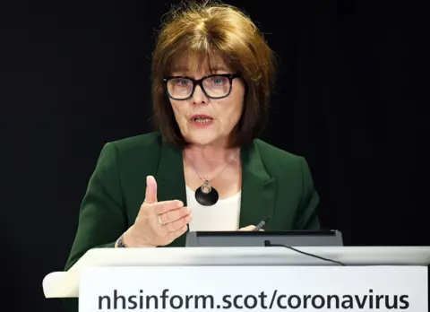 WPA POOL/Getty Images A brown haired woman wearing dark framed glasses, a white blouse and green jacket, standing at a lecturn with nhsinform.scot/coronovirus written on it