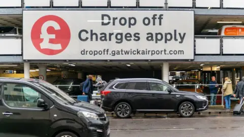 An external shot showing the drop off area at Gatwick Airport where there are two cars parked and various people getting out of them and on the pavement. There is a large sign above which reads "drop-off charges apply" and the website dropoff.gatwickairport.com