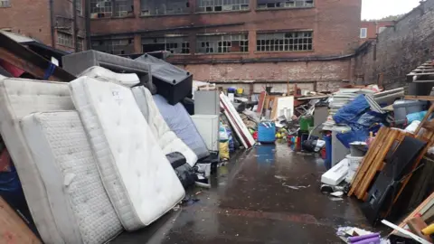 An outdoor yard filled with large quantities of discarded waste and bulky household items sits in front of a derelict multi-storey red-brick industrial building. In the foreground, several stained mattresses are stacked upright alongside upholstered furniture, cabinets, shelving, timber, plastic containers, and black rubbish bags. 