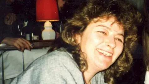 Deborah Steel has curly shoulder length hair and is smiling at the camera while leaning over lightly. She wears a light grey top. Behind her someone is smoking a cigarette and there is a white lamp with a red lamp shade.