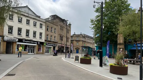 A pedestrianised shopping street with planters and an ornamental memorial on the right. Superdrug is visible on the left.  The are also planters of flowers and several pedestrians are on the street. 