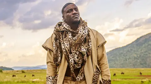 Konvict Kulture Akon is looking to the left upwards. He is wearing a cream coloured jacket with leopard printed thin scarf wrapped around his neck, going all the way to his midriff. The background is a mountain landscape with green grass, with cloudy sky illuminated by a hint of sunshine.