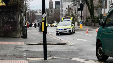 BBC A police car is positioned at an angle blocking North Hill, Plymouth. A police officer is positioning road signs in the road. Orange cones block the other lane.