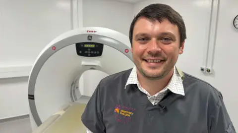 Phillip Norton/BBC Josh Smith, a man with neat dark-brown hair and stubble smiles as he stands in a white-walled veterinary surgery in front of a large CT scanner. He is wearing grey vet scrubs with a badge reading: "Aldgate Veterinary Practice".