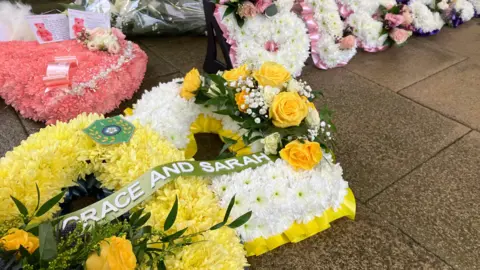 Floral displays laid on the pavement. Yellow and white flowers are side by side with a ribbon across the middle that says Grace and Sarah.