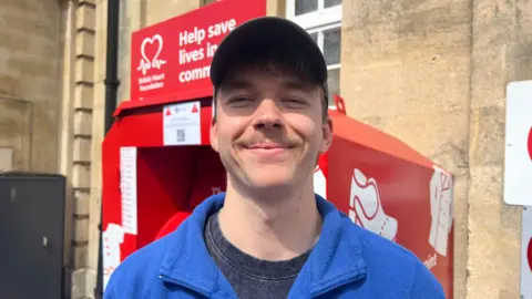 Man in blue zip top wearing a baseball hat, smiling and standing in front of a red British Heart Foundation donation bin