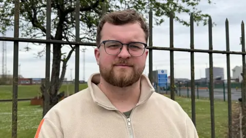 BBC/Grace McGrory Thomas Smith stands outdoors in front of tall metal railings and a grassy area, facing the camera. He wears glasses and a beige fleece. Industrial buildings, signs, and a roadway are visible in the background under an overcast sky.