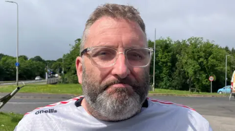 Gareth Peoples has short brown hair and a beard. He is wearing a white and red Derry GAA top and transparent and black-rimmed glasses. He is standing near a road