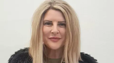 Councillor Maxine Clark - a woman with long blonde hair and wearing a black feathery top with a green shirt - smiling as she stands in front of a white wall.