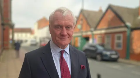 BBC Graham Stuart MP - a man with white hair - staring into the camera smiling. He is wearing a grey suit with a red tie and a light blue shirt. He is standing in Hedon town centre.