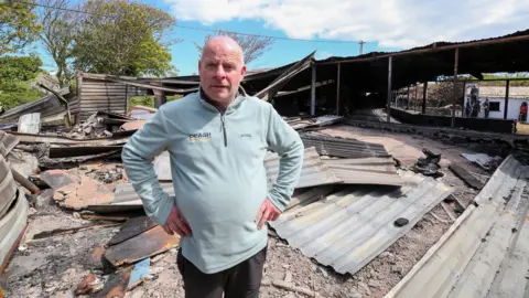 Lorcan Doherty A man wearing a blue jumper stands in the wreckage of a fire. he has his hands on his hips and stares into the camera. There is metal sheeting lying on the ground.