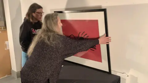 IOM ARTS COUNCIL Carola rush, who has long hair and is wearing a brown jumper, placing a red painting against a wall while Suzie Walker looks on.