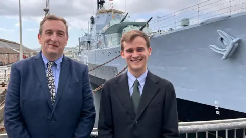 Paul Barnard and Keir Mather stand beside a large grey historic warship at Chatham Historic Dockyard. Both are wearing dark jackets and ties and are positioned in the foreground. The ship's hull, anchor and deck structures fill the background, with dock buildings visible to the left.