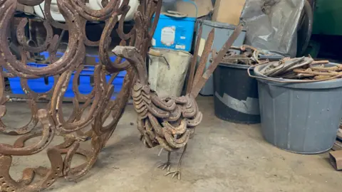 In the foreground is a sculpture of a pheasant made out of horseshoes - behind it you can see buckets of horseshoes ready to be remoulded into artwork. Next to the pheasant is a large circular structure, again made out of horseshoes. 