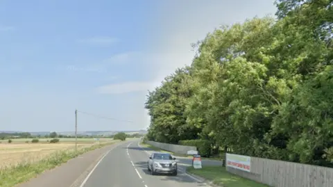 The A64 in North Yorkshire with open fields on the left of the rad and on the right the main entrance to the Merry Lees Caravan Park near Scarborough. The site has a wooden fence and large trees.