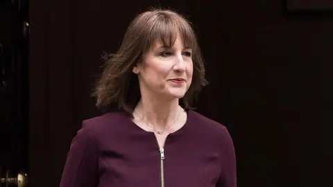 Rachel Reeves is dressed in an aubergine suit which zips up. She is pictured from the torso up and has brown mid length hair, with a fringe and wears a berry coloured lipstick. She has a small necklace and a neu. 