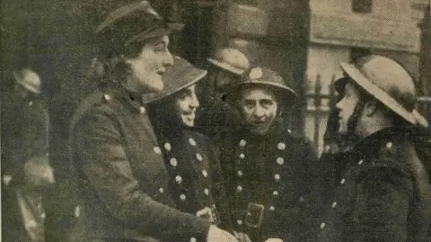 London Fire Brigade Black-and-white photo of Gillian "Bobby" Tanner in uniform during World War Two. She is smiling and wearing a peaked cap with the Auxiliary Fire Service badge. Three other uniformed men are visible beside her.
