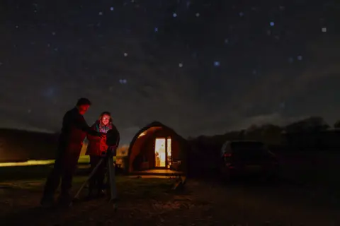 BBC/Thomas Winstone Dafydd Wyn Morgan and Siân Harrison in front of a lit up hut. They are lit by red as they set up a smart telescope. Stars are out of focus in the background. 