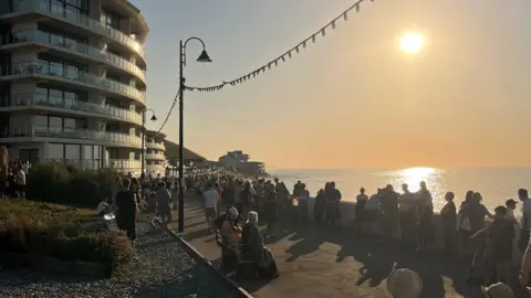 Mikey Shez People gathered along a seaside promenade (Westward Ho!) watching the sunset, with apartment buildings on one side and the sea on the other.