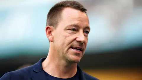 PA Media John Terry walking in front of a blurred background. He has short brown hair and is wearing a navy top.