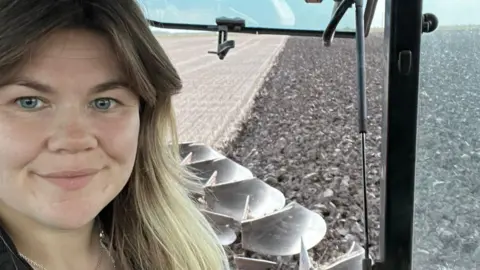 Lucy Munns Lucy, a young woman with blonde hair and blue eyes, sitting in a tractor. A wheat field is visible in the background