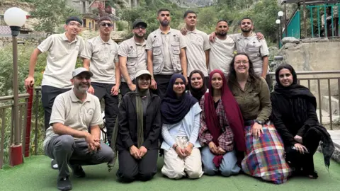A group photo of approximately 14 people posing outdoors on a terrace in a mountain valley. Snow-capped peaks rise in the background above trees and buildings, with a visible sign reading “Zipline” on a nearby structure.