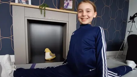 Amelia doing the splits. She's wearing a navy tracksuit and white trainers.