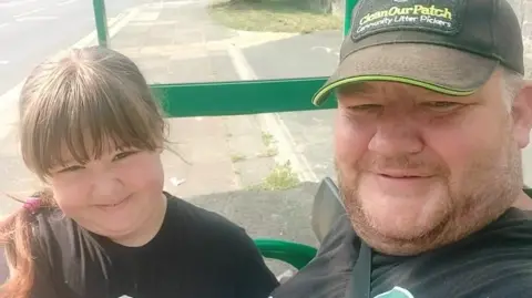 Jamie Scarlett and Jamie sat together at a bus stop. They are both smiling at the camera. 