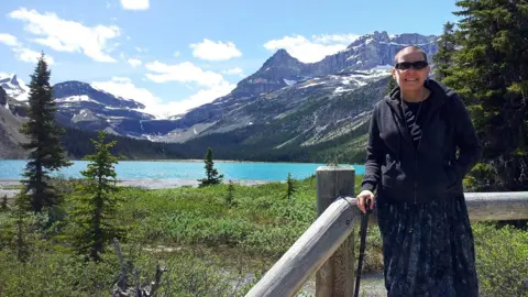 Cindy Baldassi Cindy Baldassi is in the foreground wearing sunglasses and a dark hoody and navy blue dress. She is looking at the camera and behind her is a beautiful blue lake and towering mountain peaks with snow on top of them.