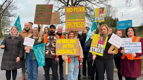 Clare Worden/BBC Group shot of striking workers outside of Duke of Lancaster school in Fakenham. They have a range of brightly coloured signs saying they are striking because of concerns about how seriously the school takes violence against staff. Some of the group have high visibility orange tabards. 