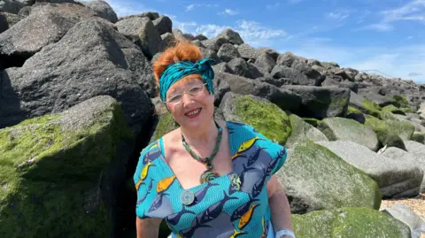 PHIL HARRISON/BBC A middle-aged woman with red hair wears a colourful turquoise sun dress decorated with a fish print design, along with a matching headscarf, as she sits on some grey rocks near the coastline.